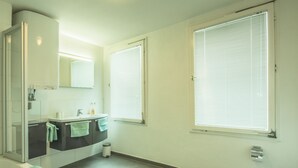 Ein Badezimmer mit Badewanne, Toilette, Waschbecken und zwei Fenstern mit Jalousien.