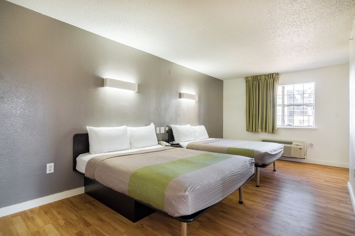 A hotel room with two beds, a window with curtains, a wall-mounted air conditioner, and a light switch on the wall.