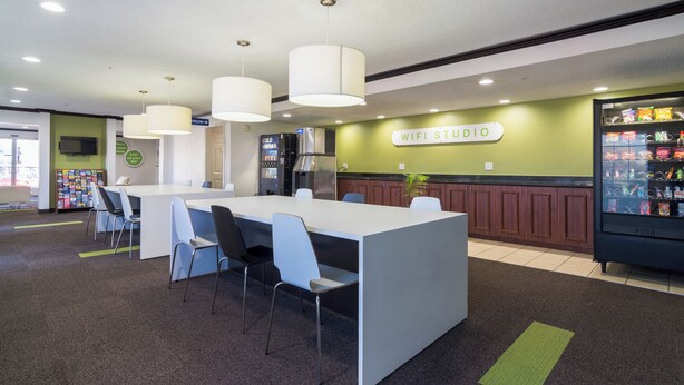 A modern indoor space with a white reception desk, multiple chairs, a vending machine, and a sign indicating a WiFi studio. - Everstay Living Extended Stay (Plano)