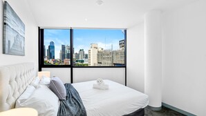 Une chambre d’hôtel avec un lit, une table de chevet, une lampe, une fenêtre donnant sur la ville et un tableau au mur.