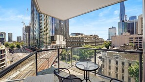 Un balcon avec une table et des chaises, donnant sur un paysage urbain composé de bâtiments modernes et historiques, d’une voie ferrée et d’un ciel dégagé.