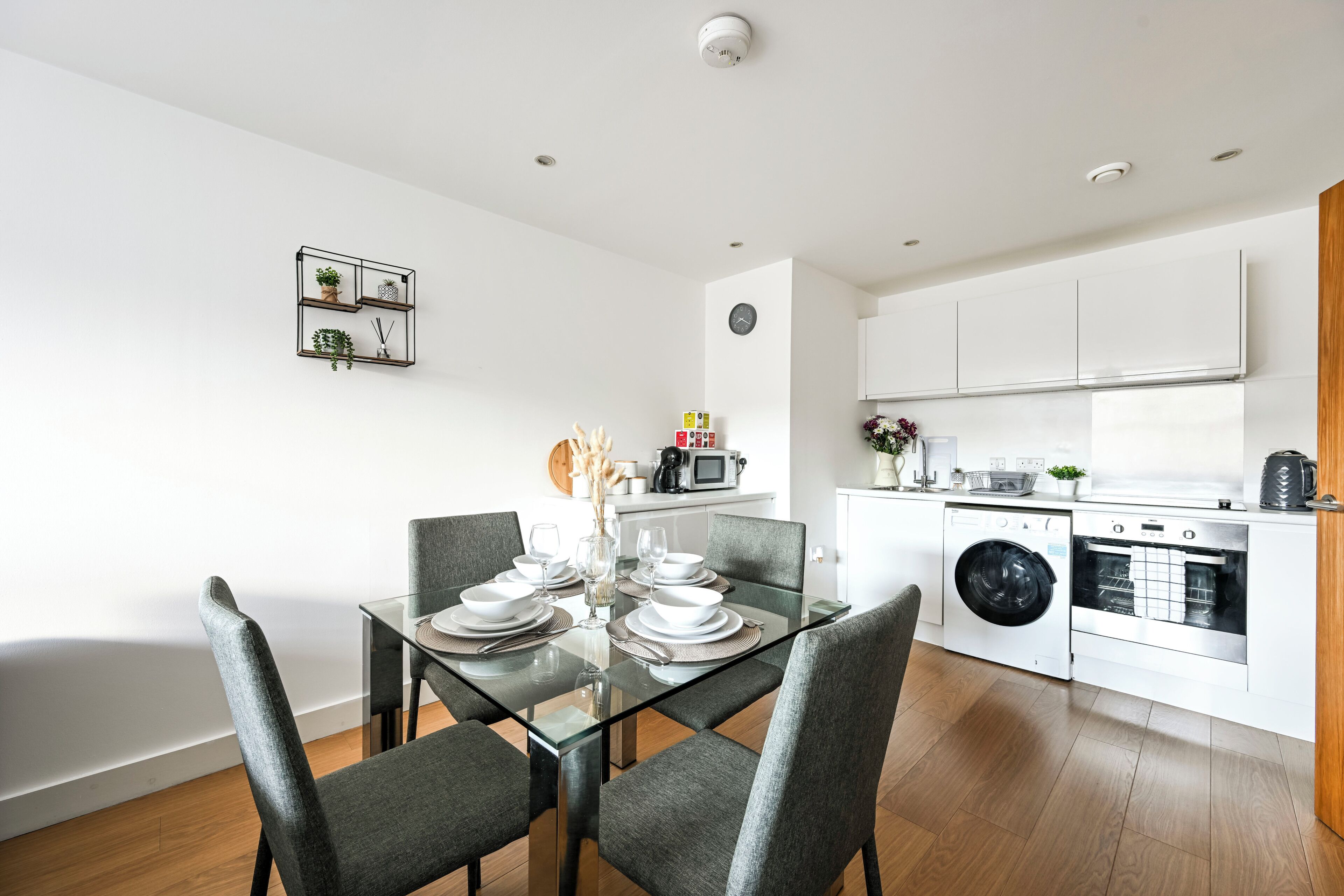 A modern kitchen with white cabinets, a washing machine, and a dining area with a glass table and grey chairs.