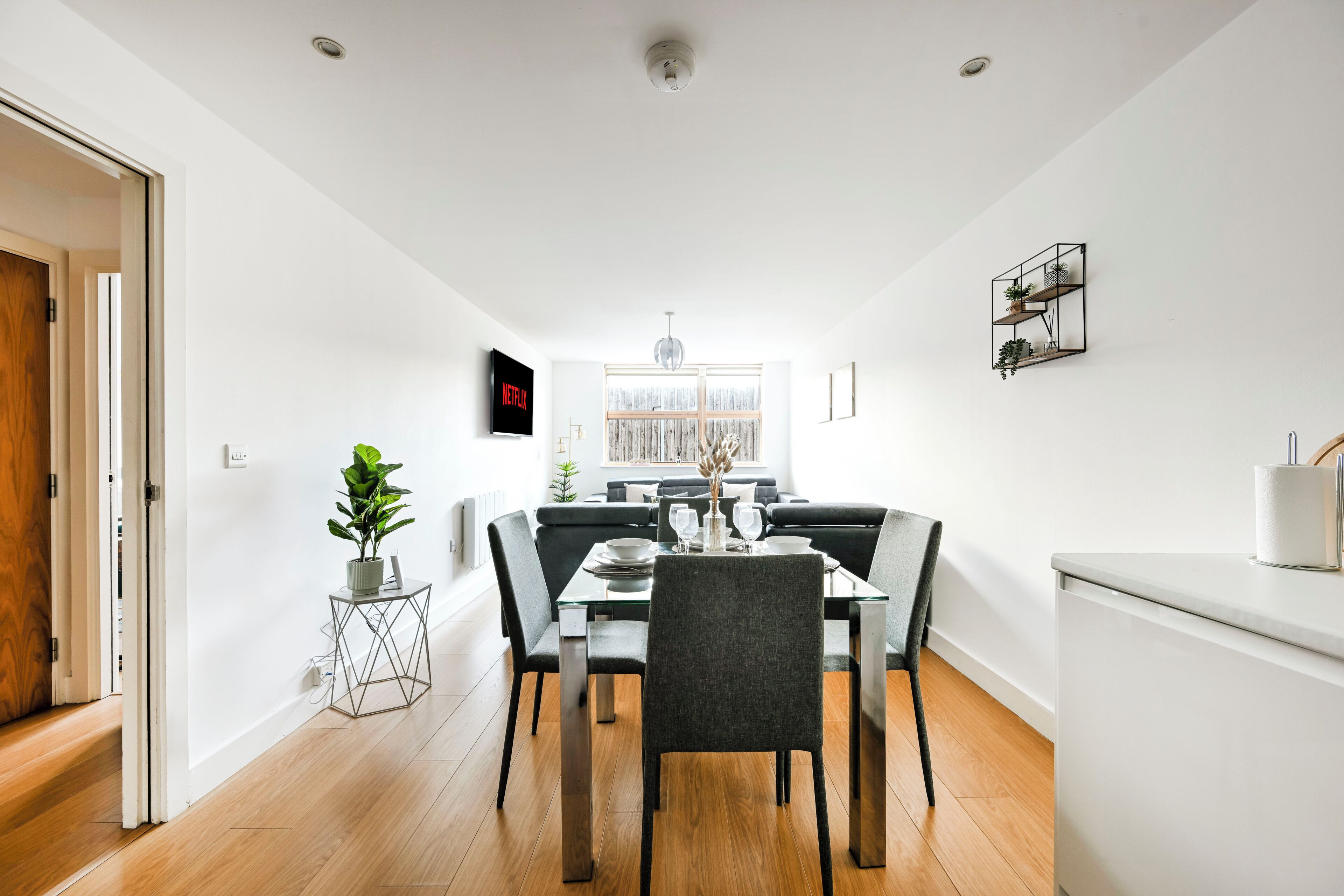 Un comedor moderno con una mesa de vidrio, sillas grises, una planta en maceta y un televisor montado en la pared.