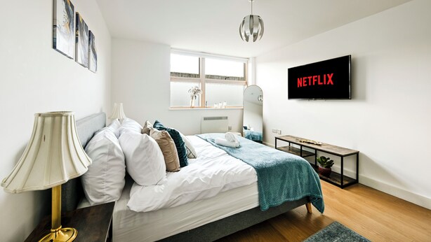 Et moderne soveværelse med seng, sengebord, lampe og et vægmonteret fjernsyn, der viser 'NETFLIX'.