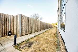 A fenced yard with a concrete path, a black bollard, and a building with large windows. - Modern London Apartment with Secure Parking. Sleeps up to 6. (London)