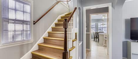 Una escalera con escalones y barandilla de madera, que conduce a una habitación con zona de comedor y televisor.