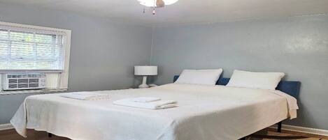 A bedroom with a bed, a ceiling fan, a window with blinds, and a wall-mounted air conditioning unit.