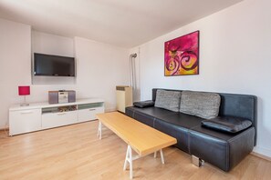 A modern living room with a sofa, a wooden bench, a flat-screen TV, a radiator, and a colorful abstract painting. - Haus Spiegel - Holiday apartment with roof terrace - Mediterran 3 (Lindau (Bodensee))