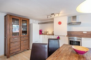 A kitchen with a wooden cabinet, a dining table with chairs, a pink bowl, and a stove. - Haus Spiegel - Holiday apartment with roof terrace - Mediterran 3 (Lindau (Bodensee))