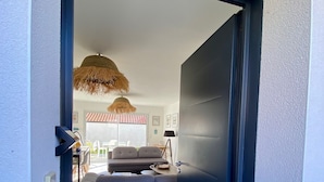 En öppen dörr leder in till ett vardagsrum med en soffa, ett litet bord och en hängande lampa.