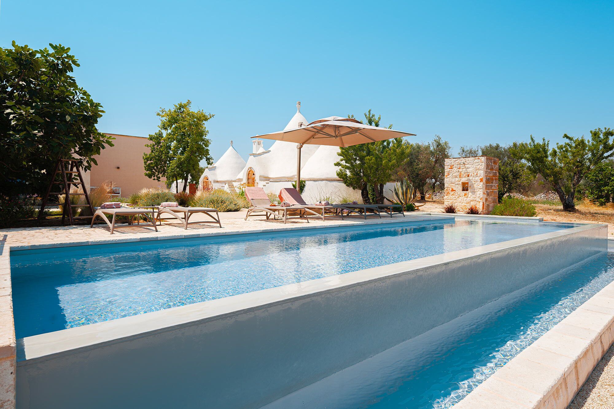 Piscina con sillas de descanso y sombrillas, rodeada de árboles y un edificio con cúpulas blancas.