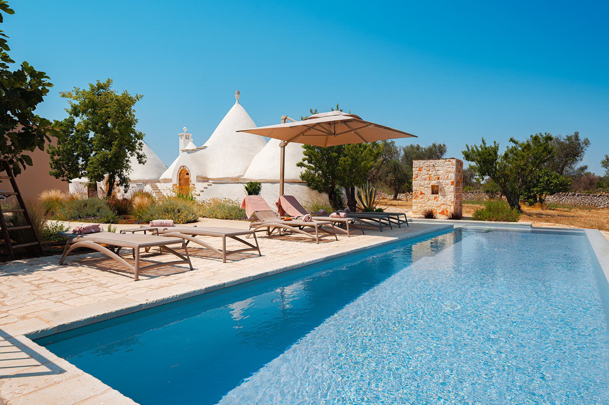 A pool area with lounge chairs and umbrellas, surrounded by white buildings and trees.