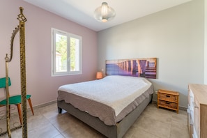 Ein Schlafzimmer mit Bett, Fenster, einem Gemälde, einer Lampe, einem Sessel und einer Kommode.