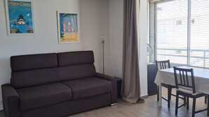 Ein Zimmer mit einem dunkelbraunen Sofa, einem Esstisch mit Stühlen, einer Stehlampe und zwei Bildern an der Wand.