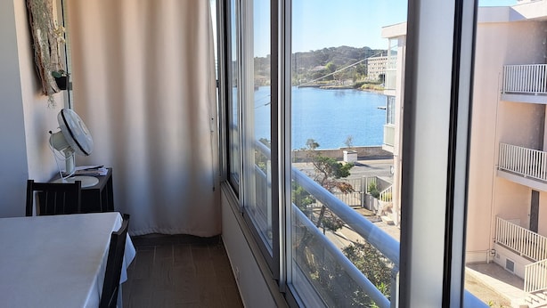 Ein Balkon mit Blick auf einen Fluss und Gebäude, ein Tisch, ein Stuhl und ein Ventilator.