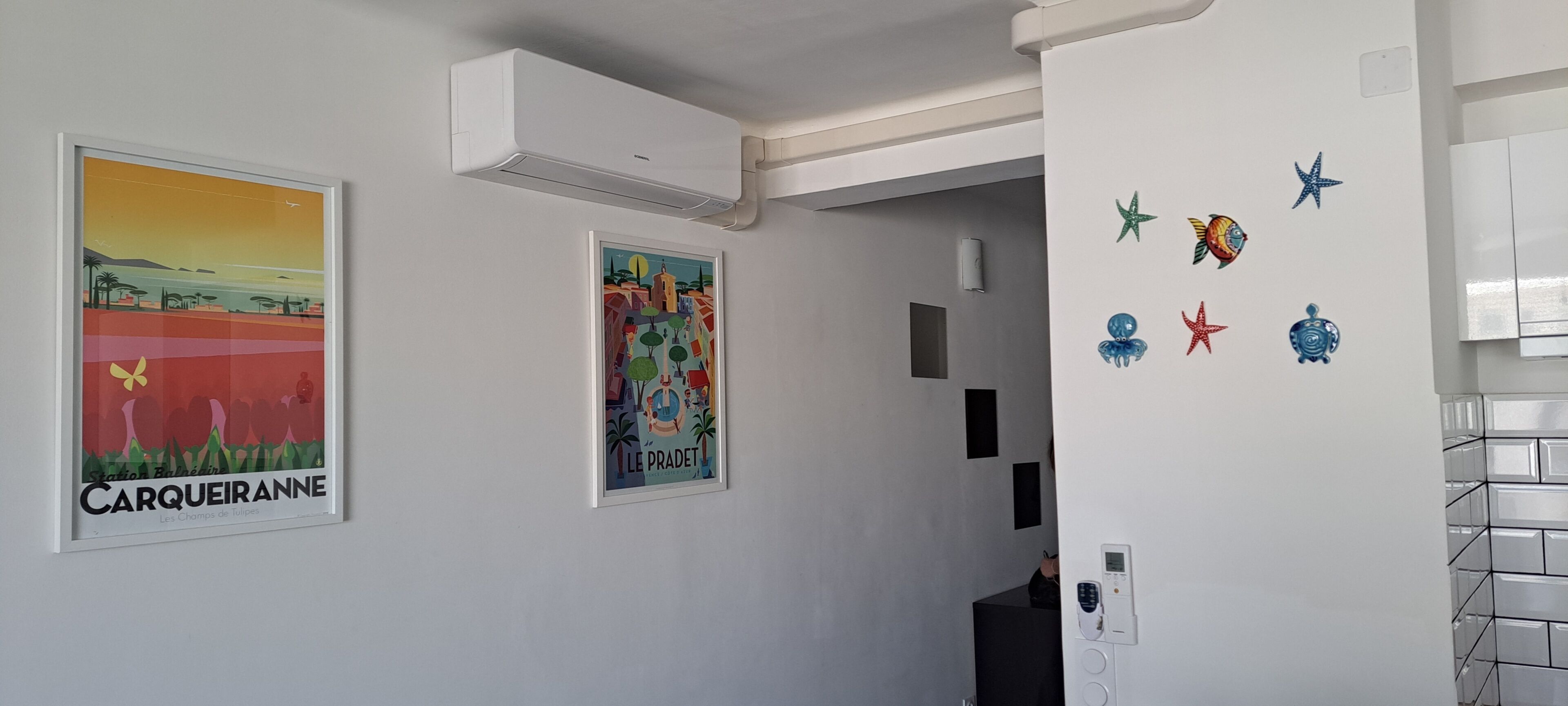 Een gang met twee ingelijste posters, een luchtverwarmingsunit aan de muur en een beveiligingspaneel.