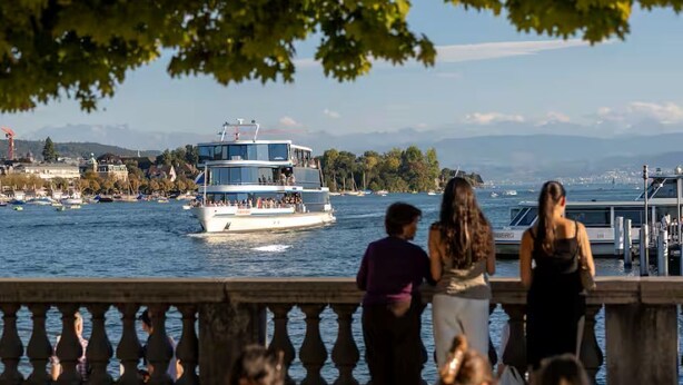 A waterfront promenade with a bench, people sitting and standing, and a boat in the background.