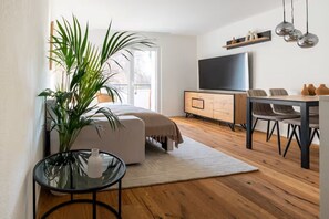 A modern living room with a sofa, a glass table, a TV, and a dining area with chairs. - 5 min to Zurich Airport, Riverside Balcony, Ergonomic Workspace, Free Parking (Opfikon)