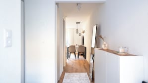 A hallway leading to a dining area with a wooden floor, white walls, and a white rug. There is a mirror on the wall and a small table with a vase and candle on the right.