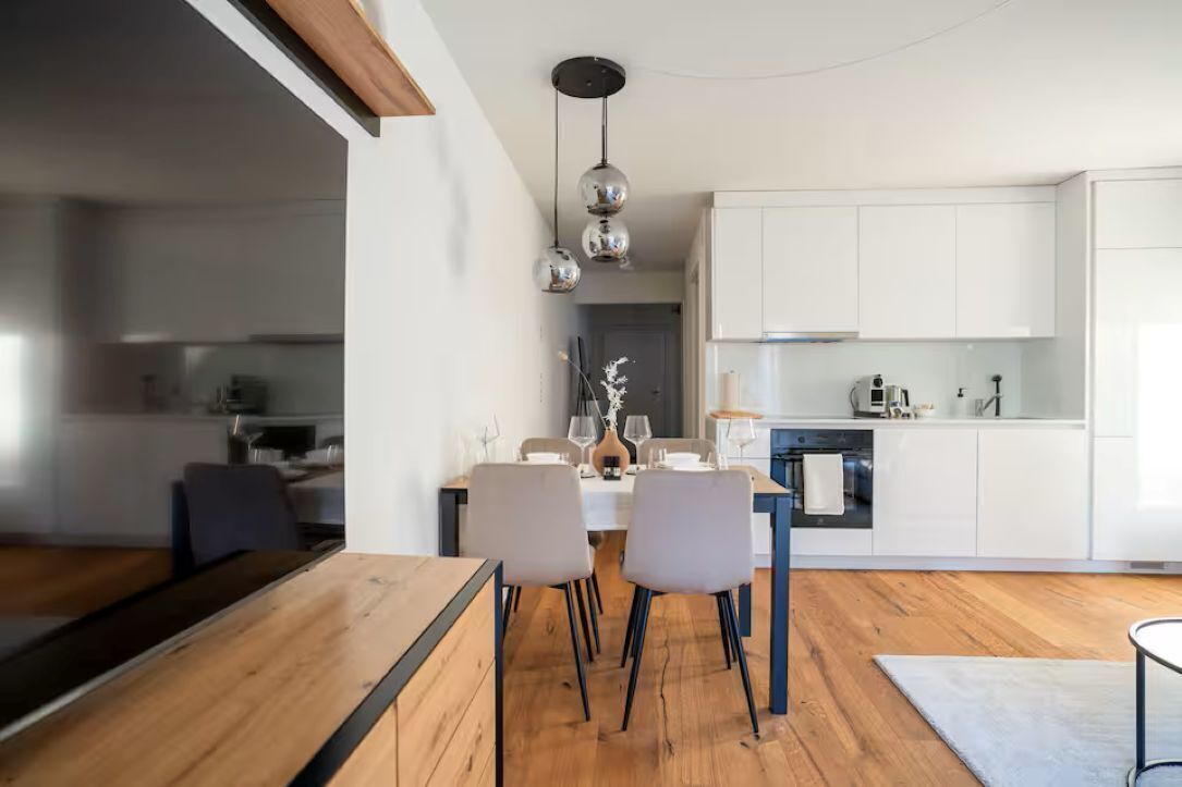 Una cucina moderna con mobili bianchi, un'area pranzo con tavolo e sedie in legno, e illuminazione a sospensione.