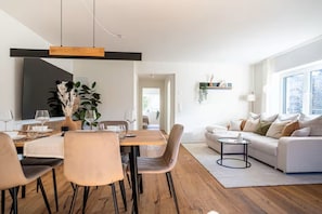 A modern living space with a dining area, a sofa, and a TV. - Spacious 2BR Apt Near Zurich Airport, Free Parking, River Access, 2 Workspaces (Opfikon)
