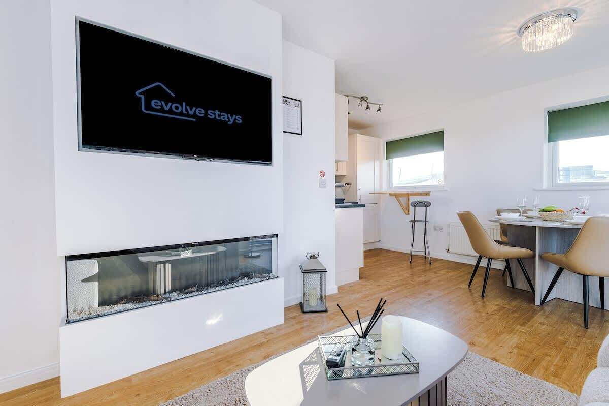 Una sala moderna con chimenea, un comedor con sillas, una cocina al fondo y un televisor mostrando 'evolve stays'.