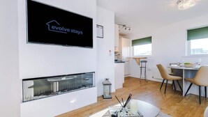 Ett modernt vardagsrum med en öppen spis, en matplats med stolar, ett kök i bakgrunden och en tv-skärm som visar programmet ”Evolve Stays”.