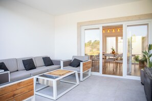 Un espace de vie moderne comprenant un canapé, une table basse et des fauteuils, un coin repas avec une table et des chaises, et une grande porte-fenêtre coulissante donnant sur un espace extérieur.