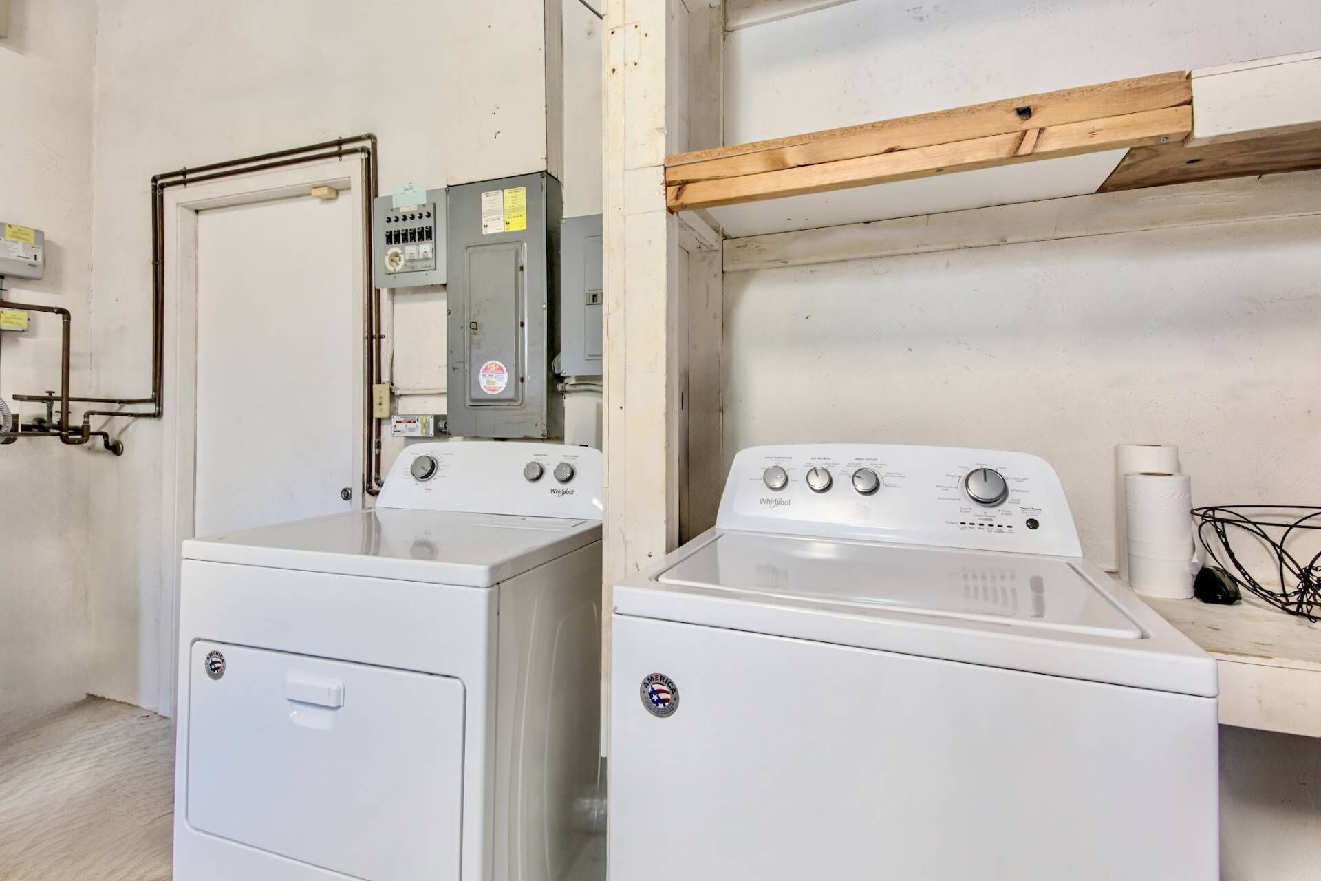 A laundry area with two Whirlpool washers, a wooden shelf, and a utility meter.