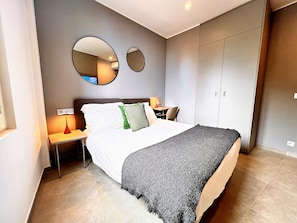 A modern bedroom with a bed, bedside tables, a lamp, a mirror, and a wardrobe. - Appartement 1B - MyResidenced'Azur (Beausoleil)
