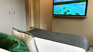 Una stanza con un letto, una televisione a parete che mostra una spiaggia tropicale e una porta che conduce in un'altra stanza.