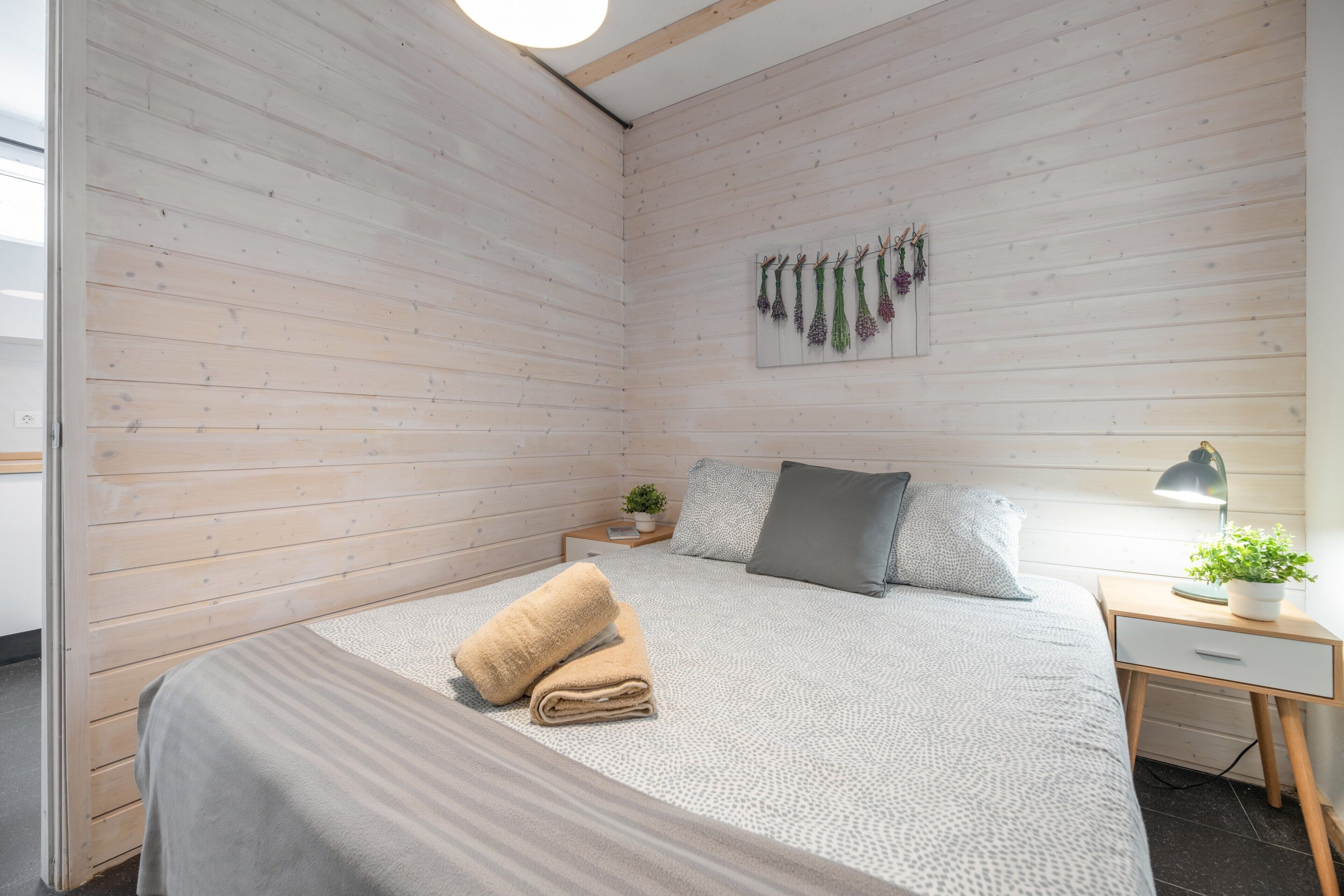 Un dormitorio con cama, mesita de noche con lámpara, planta en la pared y techo de madera.