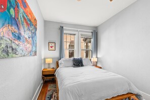 A bedroom with a bed, a nightstand, a lamp, a window with curtains, and a colorful abstract painting on the wall.