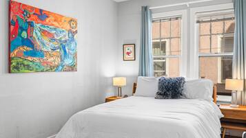 A bedroom with a bed, a nightstand, a lamp, a window with curtains, and a colorful painting on the wall.