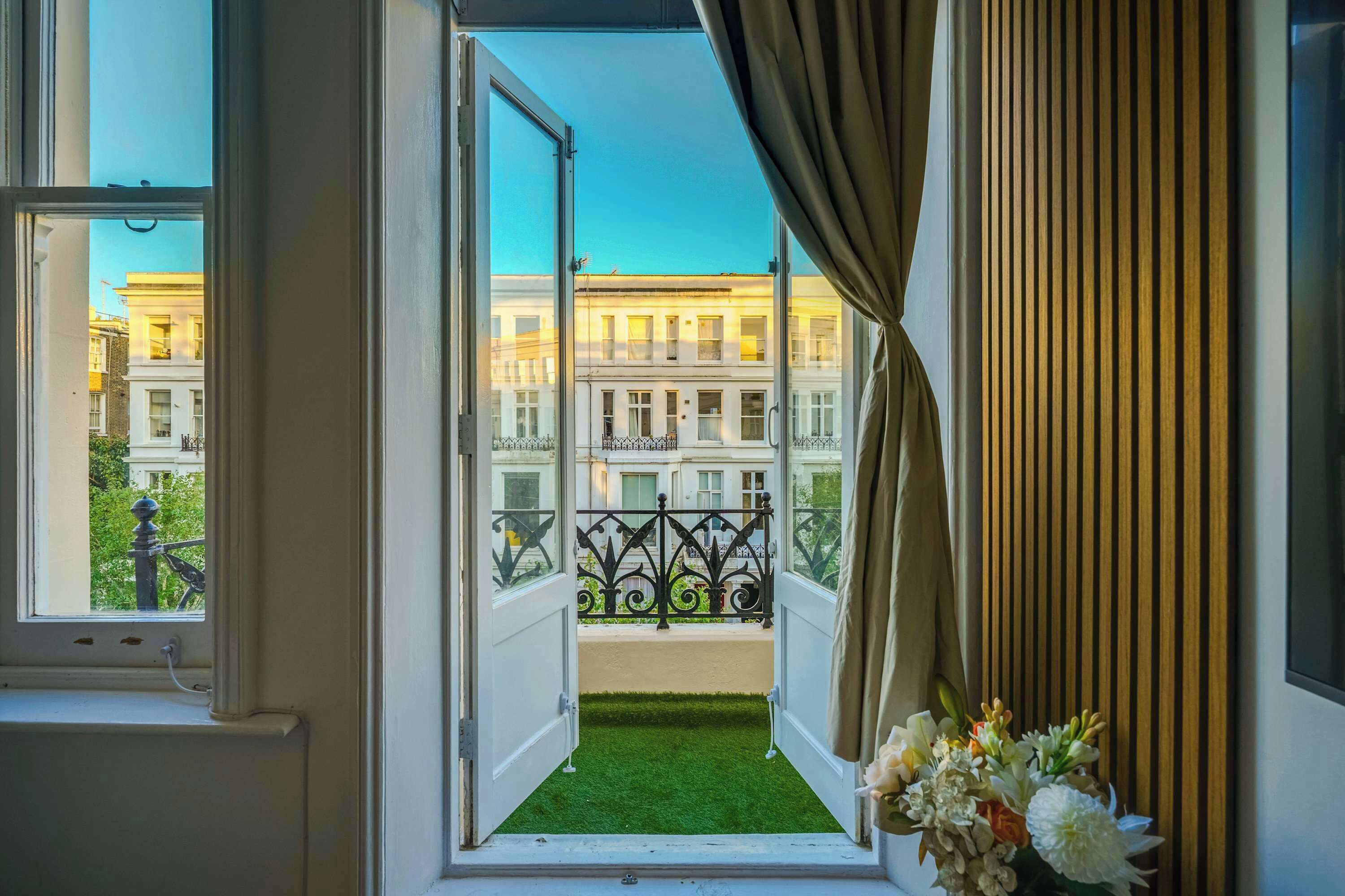 A room with a view of a building through an open window, a balcony with a metal railing, and a vase with flowers on the windowsill.