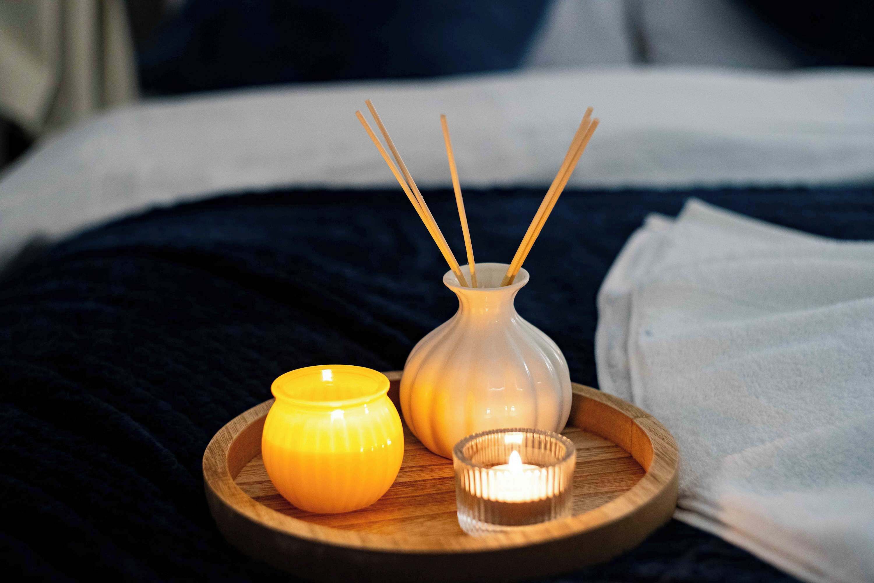 Un lit recouvert d’une couverture bleu foncé, un plateau en bois avec une bougie allumée, une bougie jaune et un porte-bougie en verre contenant une bougie allumée.