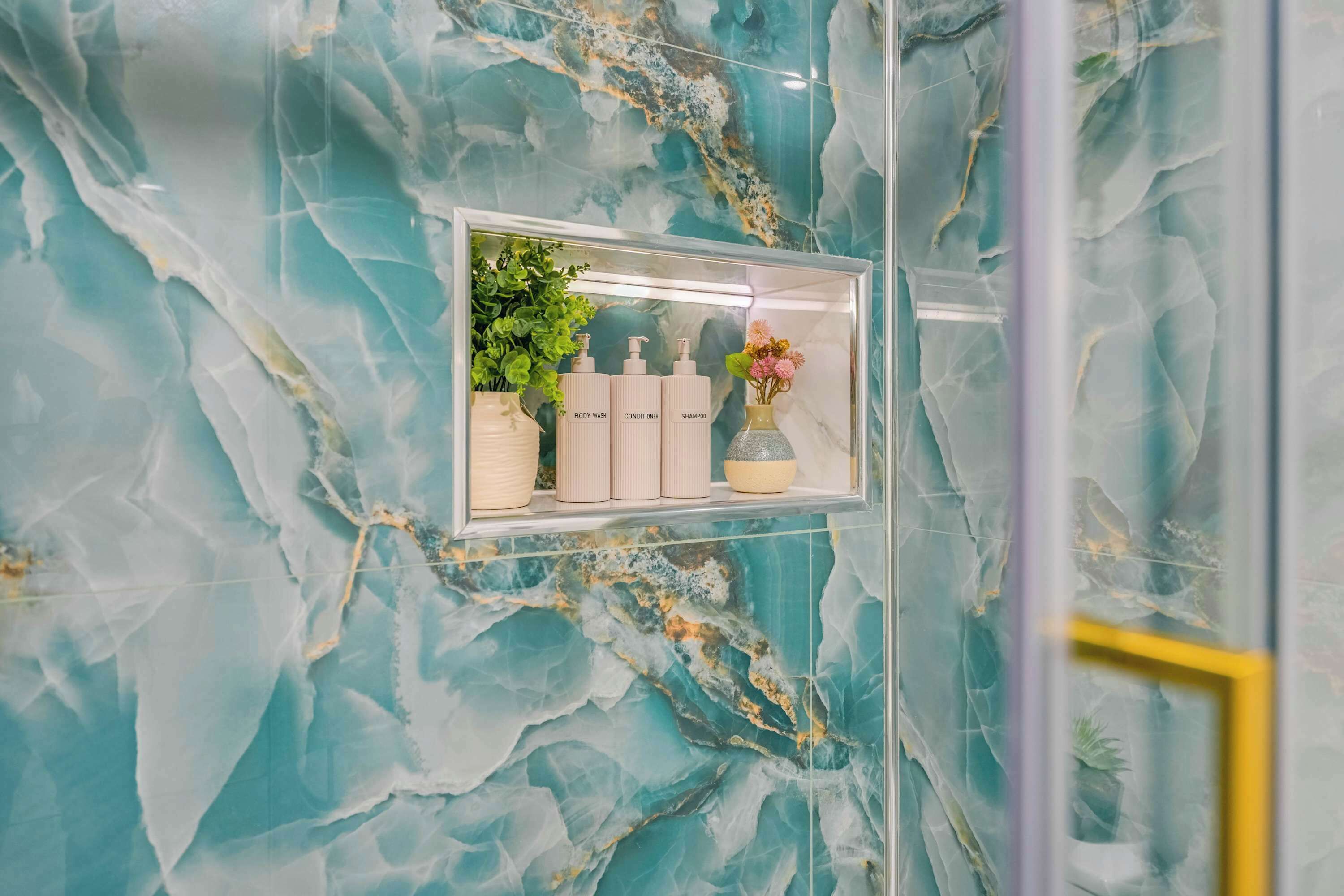 Casa de banho com parede de mármore, lavatório com divisória de vidro e produtos de higiene, e planta em vaso.