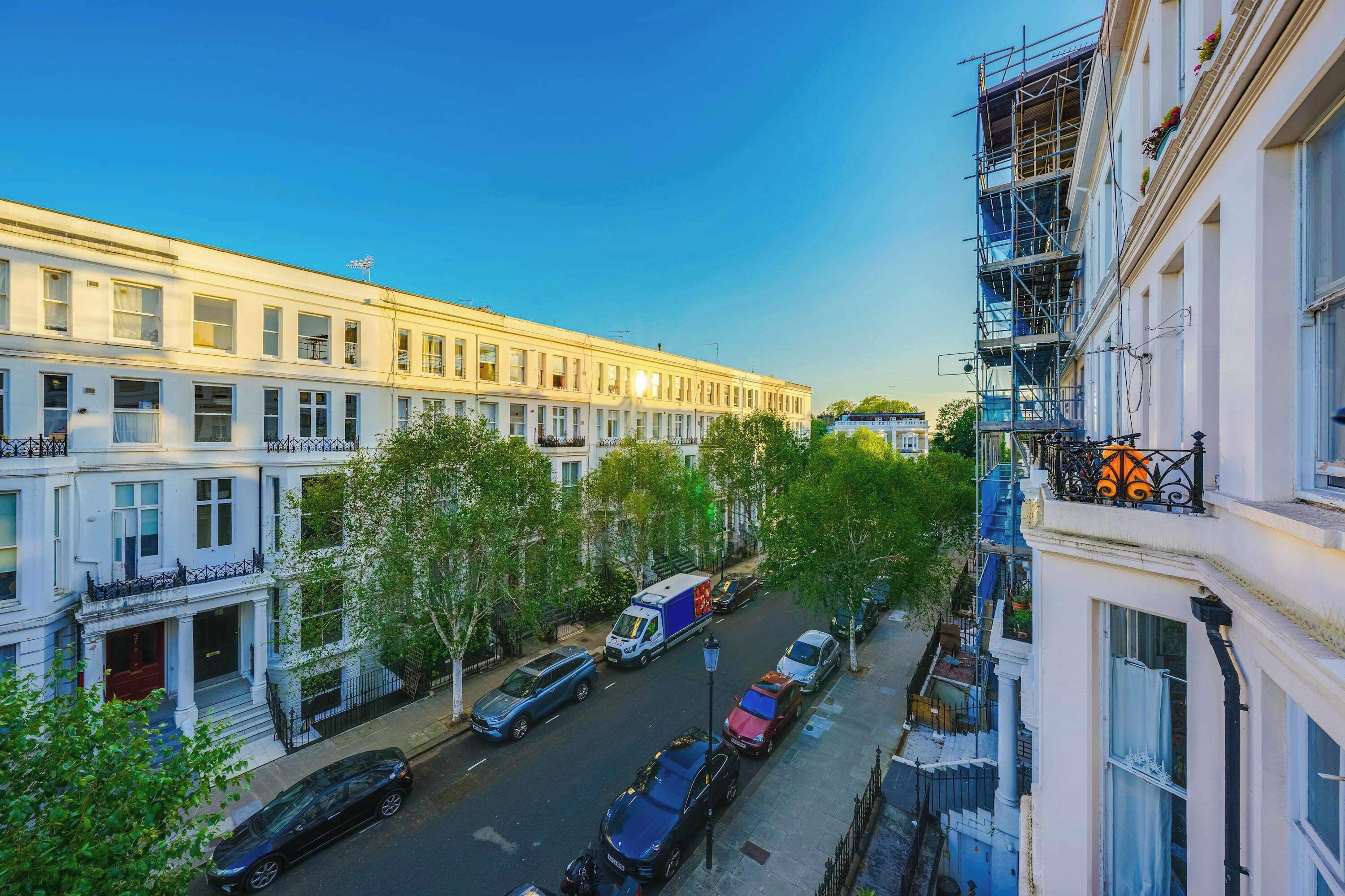 Een rij witte gebouwen met balkons, een straat met geparkeerde auto's, en een helderblauwe lucht.