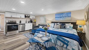 Une chambre d’hôtel compacte dotée d’une kitchenette, d’un lit recouvert d’une couvre-lit à motifs bleus et blancs, d’un coin repas avec une table et des chaises, et d’un tableau représentant des voiliers accroché au mur.