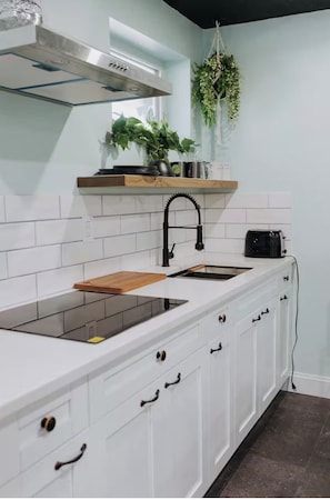 Una cocina con armarios blancos, un grifo negro, una tabla de cortar de madera y un tostador.