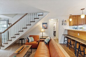 Amplia sala de estar con escaleras, un conjunto de sofás, una mesa de centro y una zona de cocina con taburetes.