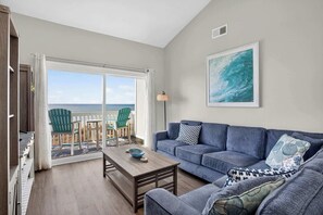 Een woonkamer met een blauwe bank, een bijzettafeltje en uitzicht op de oceaan door een glazen deur.