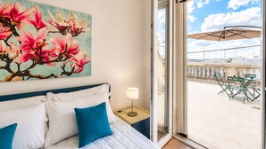 Une chambre d’hôtel avec un lit, un tableau représentant des fleurs roses, un balcon avec une table et des chaises, et une vue sur la ville.