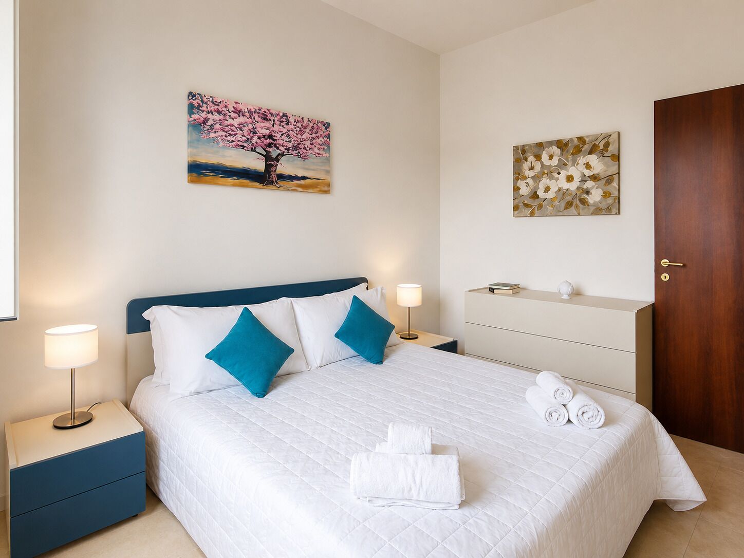 Una habitación de hotel con una cama, dos mesitas de noche, un aparador y obras de arte en las paredes.