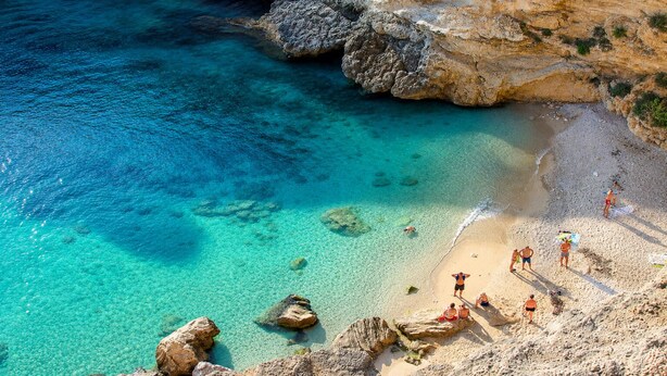 Une plage côtière bordée d’une eau turquoise et limpide, de falaises rocheuses et de personnes se prélassant au soleil.