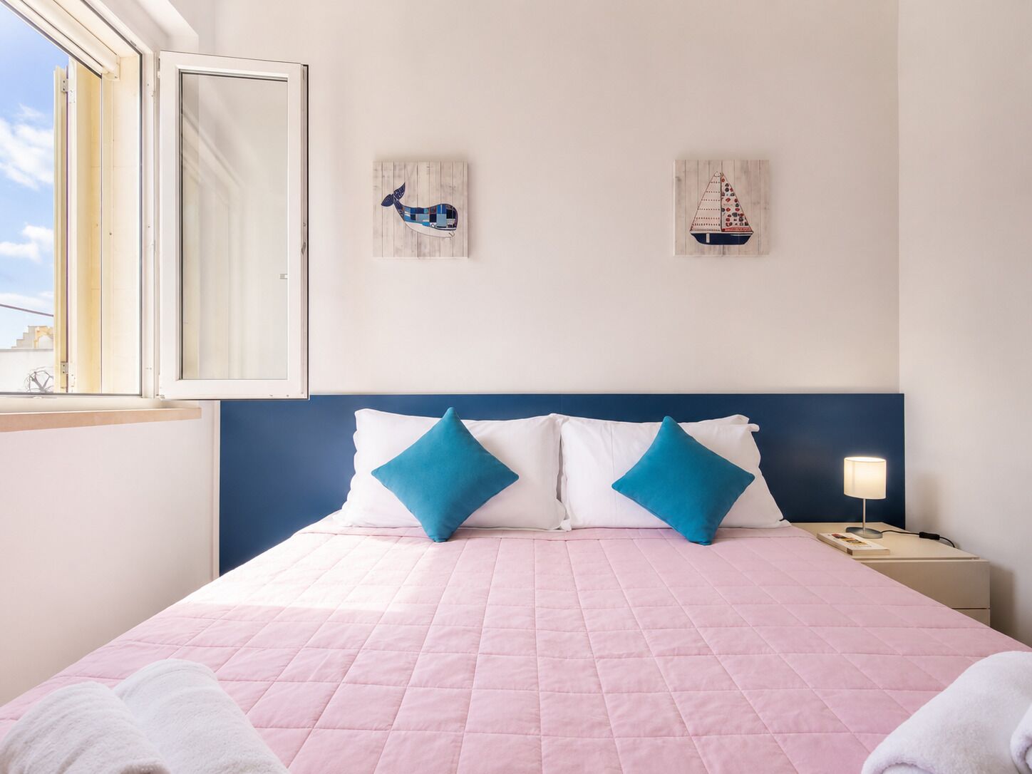 Un dormitorio con colcha acolchada rosa, dos almohadas azules, cabecera de madera, mesita de noche con lámpara y dos obras de arte en la pared.