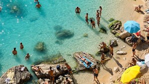 Une plage avec une eau turquoise et limpide, des formations rocheuses, et des gens qui se baignent et prennent des bains de soleil.