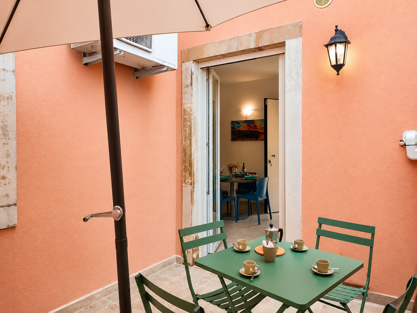 Un pequeño patio con una mesa dispuesta para dos personas, sillas, una lámpara fijada a la pared y un equipo de aire acondicionado.