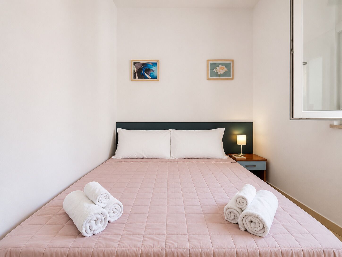 A hotel room with a bed, two pillows, a nightstand with a lamp, and two wall-mounted pictures.