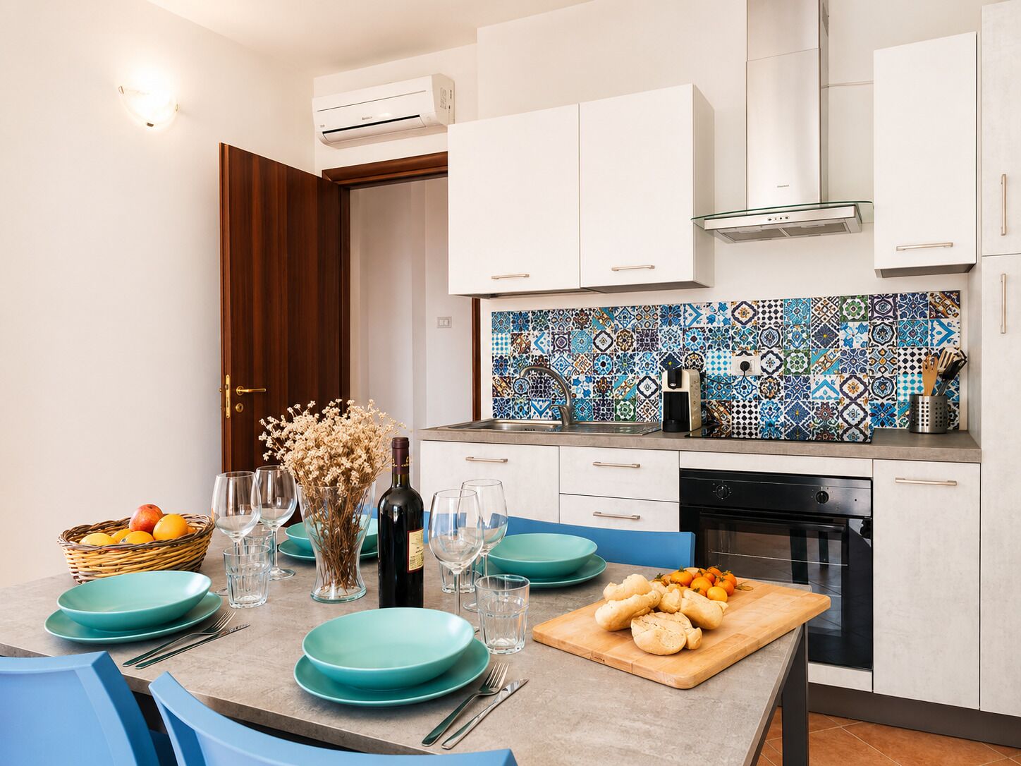A modern kitchen with white cabinets, a patterned backsplash, and a dining area with blue chairs and teal plates.